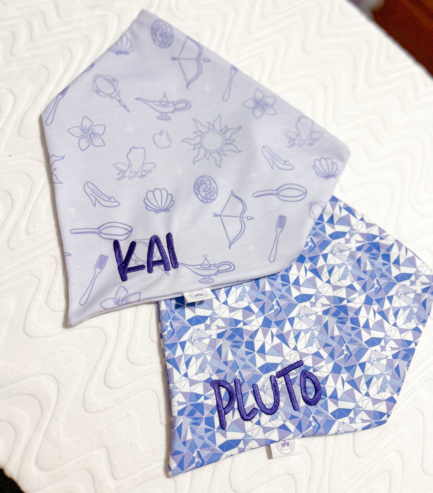 Two fabric bandanas with 'Kai' and 'Pluto' printed on them, one with a purple geometric pattern and the other with princess icons.