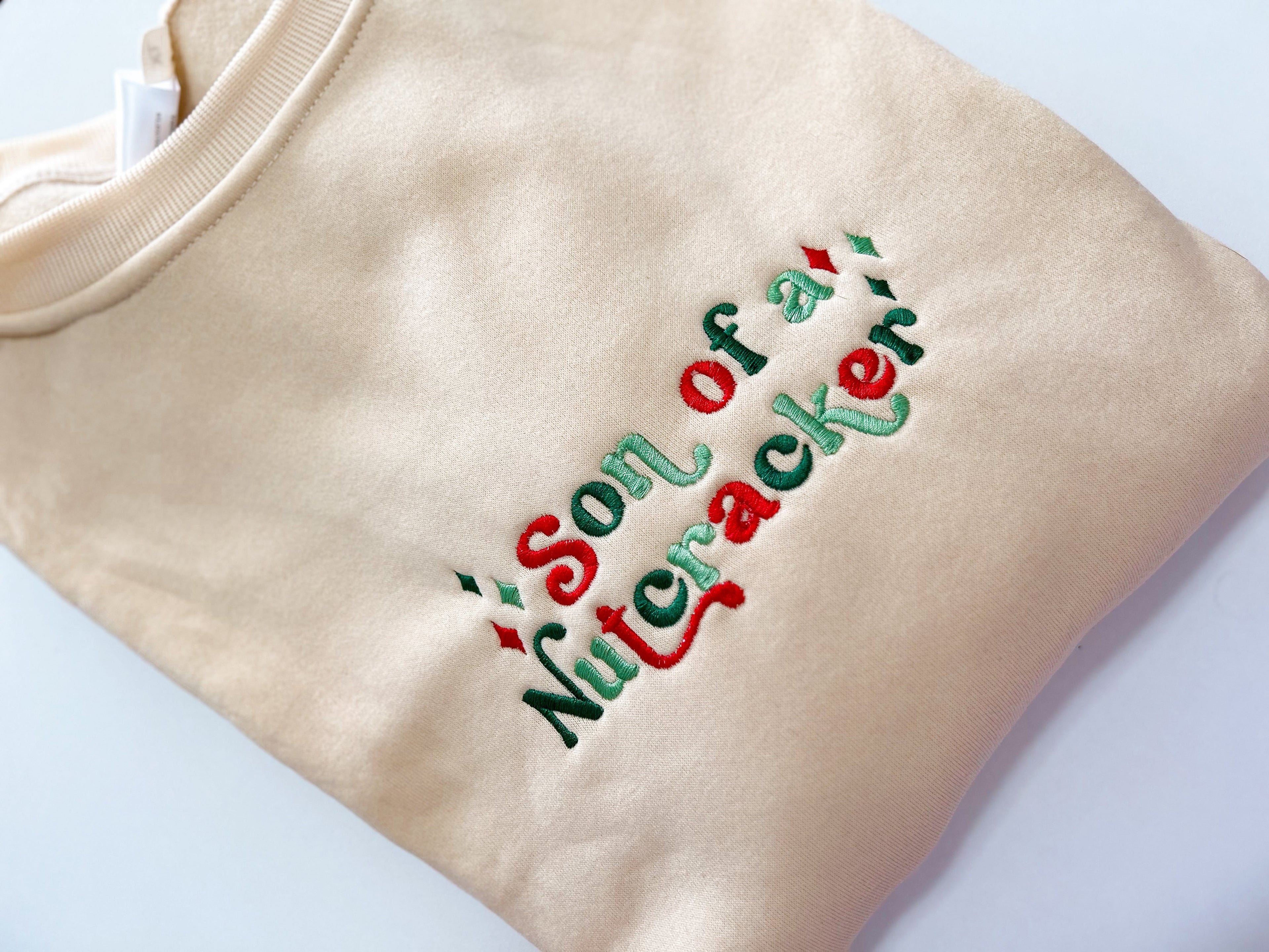 Beige sweatshirt with 'Son of a Nutcracker' text in red and green on a white background